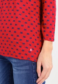 Camicia rossa con motivo di gatti blu navy, maniche a 3/4, orlo arrotondato e un piccolo dettaglio con bottone rosa nella parte inferiore a sinistra.