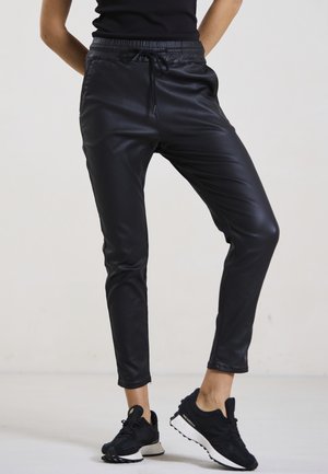 Schwarze Jogginghosen aus Kunstleder mit elastischem Bund und Kordelzug. Slim Fit mit Knöchelreißverschlüssen. Kombiniert mit schwarzen Sportschuhen.