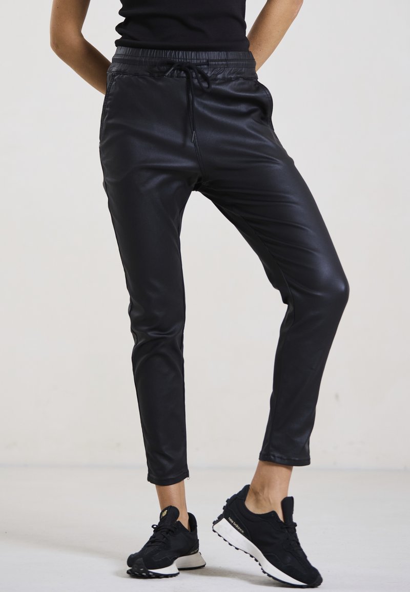 Schwarze Jogginghosen aus Kunstleder mit elastischem Bund und Kordelzug. Slim Fit mit Knöchelreißverschlüssen. Kombiniert mit schwarzen Sportschuhen.