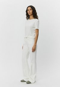 Tricou alb cu mâneci scurte, asortat cu blugi albi cu talie înaltă și evazați. Purtat cu pantofi slip-on verzi texturați. Design simplu și minimalist.