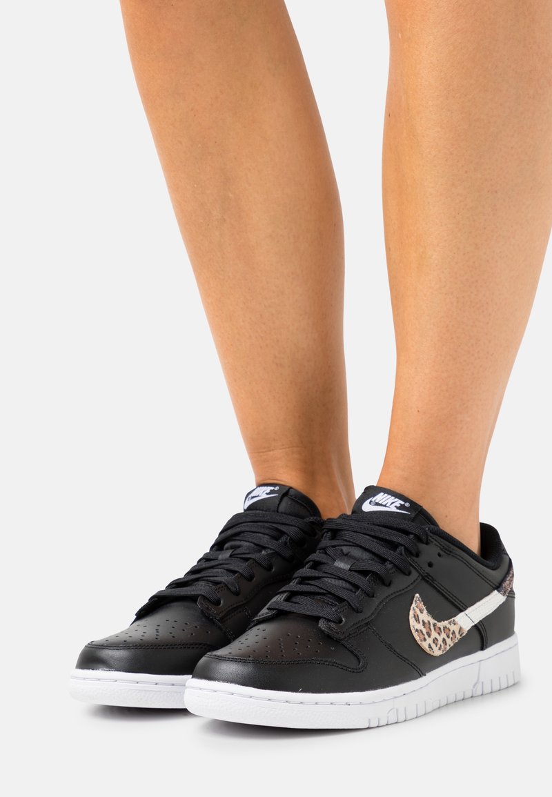 Mustat nahkaiset sneakerit valkoisella pohjalla, joissa on leopardi-kuosiset Nike-logot ja mustat nauhat. Muotoilussa on reikäpelti varvasosassa hengittävyyden parantamiseksi.