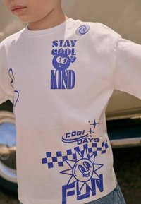 Witte katoenen T-shirt met blauwe graphics, waaronder "STAY COOL KIND," een lachende zon en geruite patronen. Regular fit met een ronde hals.