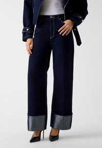 Παντελόνια από ναυτικό τζιν με φαρδιά γραμμή και αντίθετες ασημί μανσέτες. Λεπτομέρειες με ορατή ραφή και κούμπωμα με κουμπί. Συνδυασμένα με μαύρες ψηλοτάκουνες γόβες.