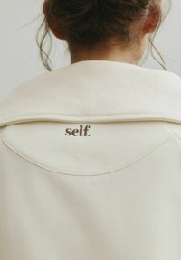 Felpa color crema con collo a costine e un ricamo sottile "self." sul retro. Texture morbida e design casual.