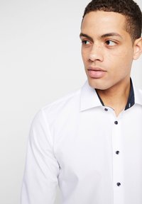 Junger Mann mit kurzen lockigen Haaren und Sommersprossen, der ein weißes Hemd mit dunklen Knöpfen trägt und auf einem einfarbigen Hintergrund seitlich schaut.