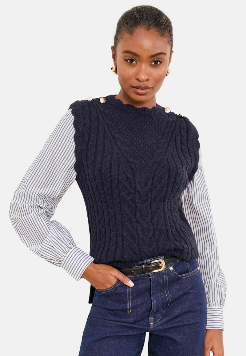 Gilet marin en maille avec motif torsadé et encolure festonnée, agrémenté de boutons dorés à l'épaule, porté sur une chemise rayée.