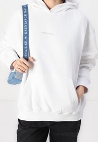 Sweat à capuche blanc avec une poche avant et des poignets côtelés ; présente un logo gris discret « PEGADOR » sur la poitrine. Associé à un jean foncé et un sac bleu.