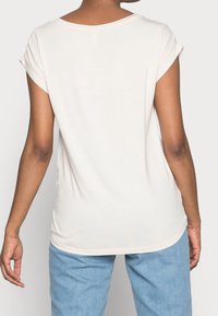Top crema de color claro, de manga corta y cuello redondo con puños enrollados, que presenta una textura suave y un corte relajado, visto desde la parte de atrás.