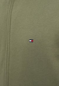 Αθλητική μπλούζα ελιάς με μικρό κεντημένο λογότυπο σε κόκκινο, άσπρο και μπλε χρώμα. Διαθέτει απαλή υφή και ορατές λεπτομέρειες ραφής.