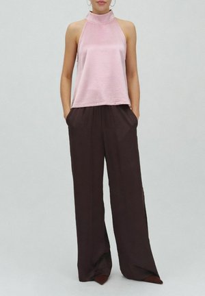 Kobieta ubrana w różowy top bez rękawów z paskiem na szyi i luźne, szerokie, ciemnobrązowe spodnie, stoi z rękami w kieszeniach.