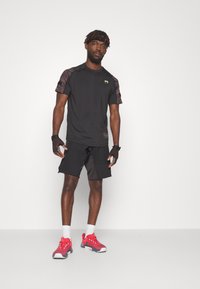 Schwarzes Sport-T-Shirt mit camo-musterhaften kurzen Ärmeln, kombiniert mit schwarzen Shorts. Rote Sneakers mit weißen Akzenten und Knöchelsocken.