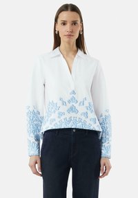 Witte blouse met blauwe borduursels op de mouwen en de onderrand, met een kraag en een knoopsluiting. De stof is glad met een lichte textuur.