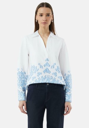 Weiße Bluse mit blauer Stickerei an den Ärmeln und am Saum, ausgestattet mit einem Kragen und einer Knopfleiste. Der Stoff ist glatt und hat eine leichte Textur.