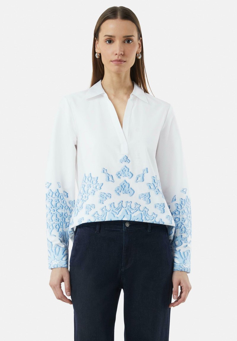 Witte blouse met blauwe borduursels op de mouwen en de onderrand, met een kraag en een knoopsluiting. De stof is glad met een lichte textuur.