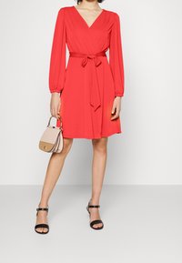 Červené šaty s dlouhými nafouklými rukávy, s páskem na zavázání a délka ke kolenům. Kombinované s černými sandály na pásky a malou béžovou kabelkou.