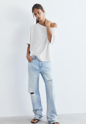 Mladá žena nosící volné bílé tričko s odhalenými rameny, světle modré roztrhané široké džíny a černé sandály, stojící před bílým pozadím.