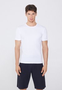 Weißes T-Shirt mit kurzen Ärmeln und rundem Halsausschnitt, aus glattem Stoff. Getragen mit dunkelblauen Shorts, vor einem neutralen Hintergrund stehend.