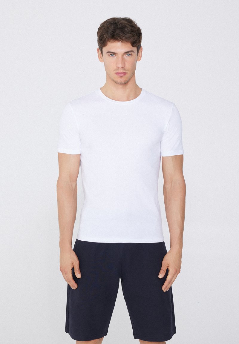 Weißes T-Shirt mit kurzen Ärmeln und rundem Halsausschnitt, aus glattem Stoff. Getragen mit dunkelblauen Shorts, vor einem neutralen Hintergrund stehend.