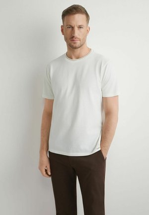 Mężczyzna o krótkich włosach, ubrany w gładką białą koszulkę i ciemnobrązowe spodnie, stoi oparty o białą ścianę, z jedną ręką w kieszeni.