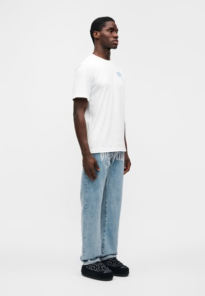 Λευκό βαμβακερό T-shirt με κοντά μανίκια, συνδυασμένο με ανοιχτό μπλε τζιν που έχει γραφικό σχέδιο σε λευκό. Το μοντέλο φοράει μαύρες κεντημένες εσπαντρίγιες.