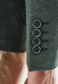 Zielona marynarka w kratę z rękawami ozdobionymi czterema ciemnymi guzikami i akcentami w postaci ściegów. Tkanina ma teksturowany splot, co dodaje głębi projektowi.