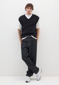 Jersey de punto negro sin mangas sobre una camisa blanca, combinado con pantalones de mezclilla oscuros y zapatillas deportivas grises. Diseño minimalista.