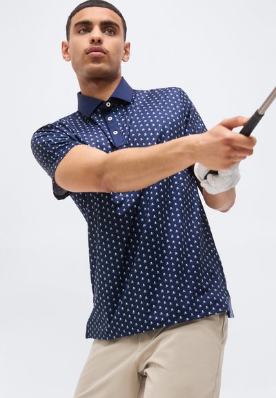 Άνδρας με μπλε πουά πόλο και μπεζ παντελόνι που κάνει κούνια με μπαστούνι γκολφ, φοράει λευκό γάντι γκολφ στο αριστερό του χέρι.