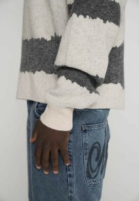 Pulover tricotat cu dungi crem și gri, material texturat, manșete cu bordură, asortat cu blugi din denim deschis la culoare, având detalii brodate pe buzunar.