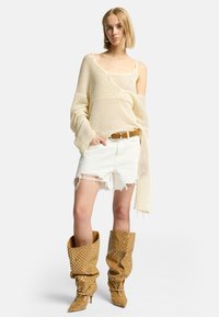 Hellbeige oversized Strickpullover mit lockeren Ärmeln, kombiniert mit weißen, ausgeblichenen Shorts und langen, spitzen karamellfarbenen Stiefeln mit weißen Punkten.
