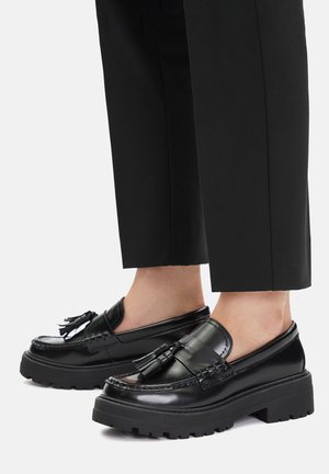 Czarne skórzane mokasyny o teksturowanej powierzchni, z grubą podeszwą i ozdobnymi frędzlami z przodu. Noszone z dopasowanymi czarnymi spodniami.