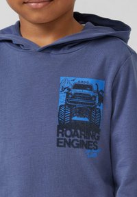 Blauwe katoenen hoodie met een voorzijdeafbeelding van een monstertruck en bergen. De tekst luidt "GROMMEND MOTOREN EN SCHERPE KLAUWEN" in zwart en blauw.