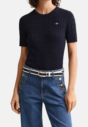 Kvinde iført en marineblå kortærmet kabelstrikket trøje og blå højtaljede jeans med guldknapper, stribet bælte med guldlås.