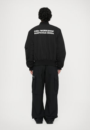 Mustaline musta bomber-jakk featuring tekst "032c WORKSHOP SUSPICIOUS MINDS" seljal, koos lõdva musta cargo-pükstega.