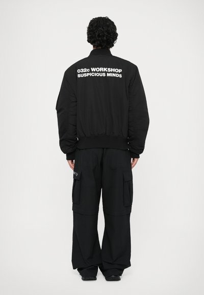 Giubbotto bomber nero con il testo "032c WORKSHOP SUSPICIOUS MINDS" sulla schiena, abbinato a pantaloni cargo neri larghi.