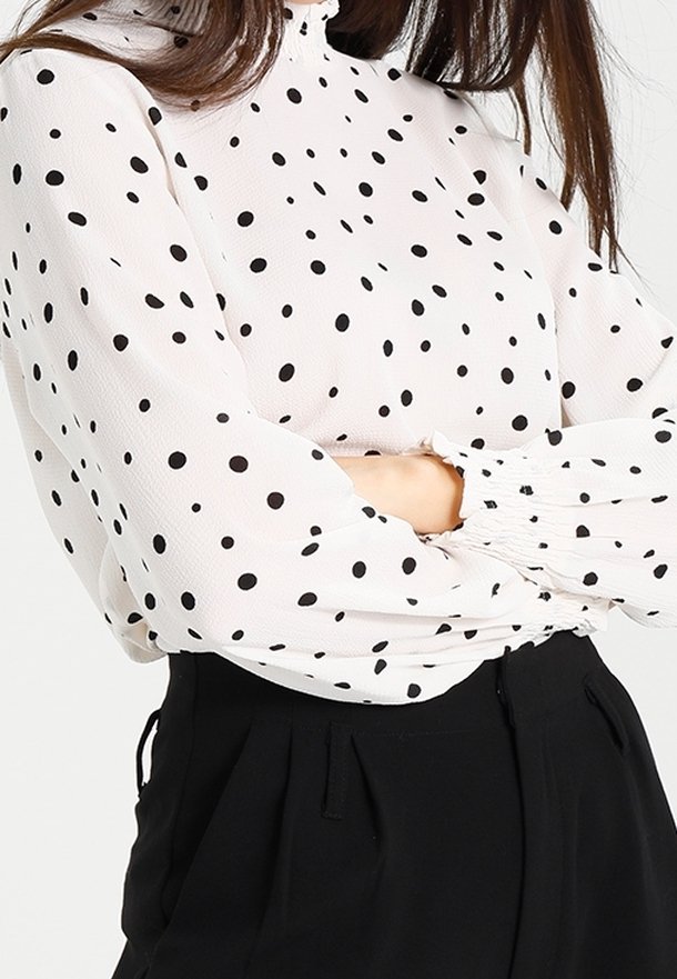 Blouse blanche à pois noirs, col montant froncé, poignets élastiqués et manches fluides, associée à un pantalon noir.