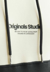 Svetlo krémová mikina s čiernym pásom a šnúrkami. Obsahuje potlačené logo: "Originals Studio" v tmavých písmenách. Strieborné kovové akcenty.