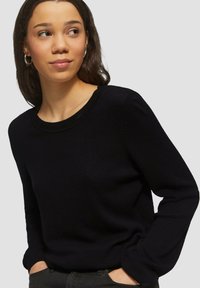 Pull en tricot noir avec un motif texturé, col rond et manches longues. Tissu léger, coupe ajustée, et il présente des détails minimaux.