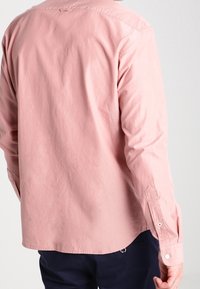 Ljusrosa långärmad skjorta med knappkrage, manschetter med en knapp och tyg med mjuk textur; har en rundad fåll och subtila sömmar.