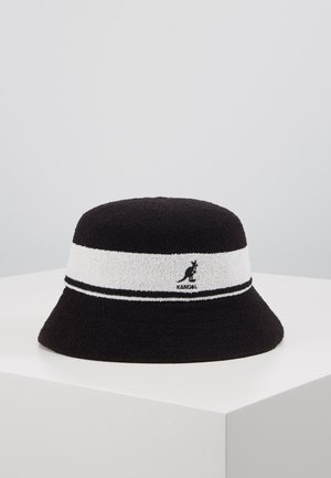 Musta ämpärilakki, jossa valkoinen nauha ja kengurun logo sekä "Kangol"-teksti, esillä valkoisella jalustalla yksinkertaista taustaa vasten.