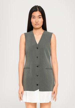 Woman with long straight black hair wearing sleeveless gray pinstriped buttoned vest dress with white ruffle hem.