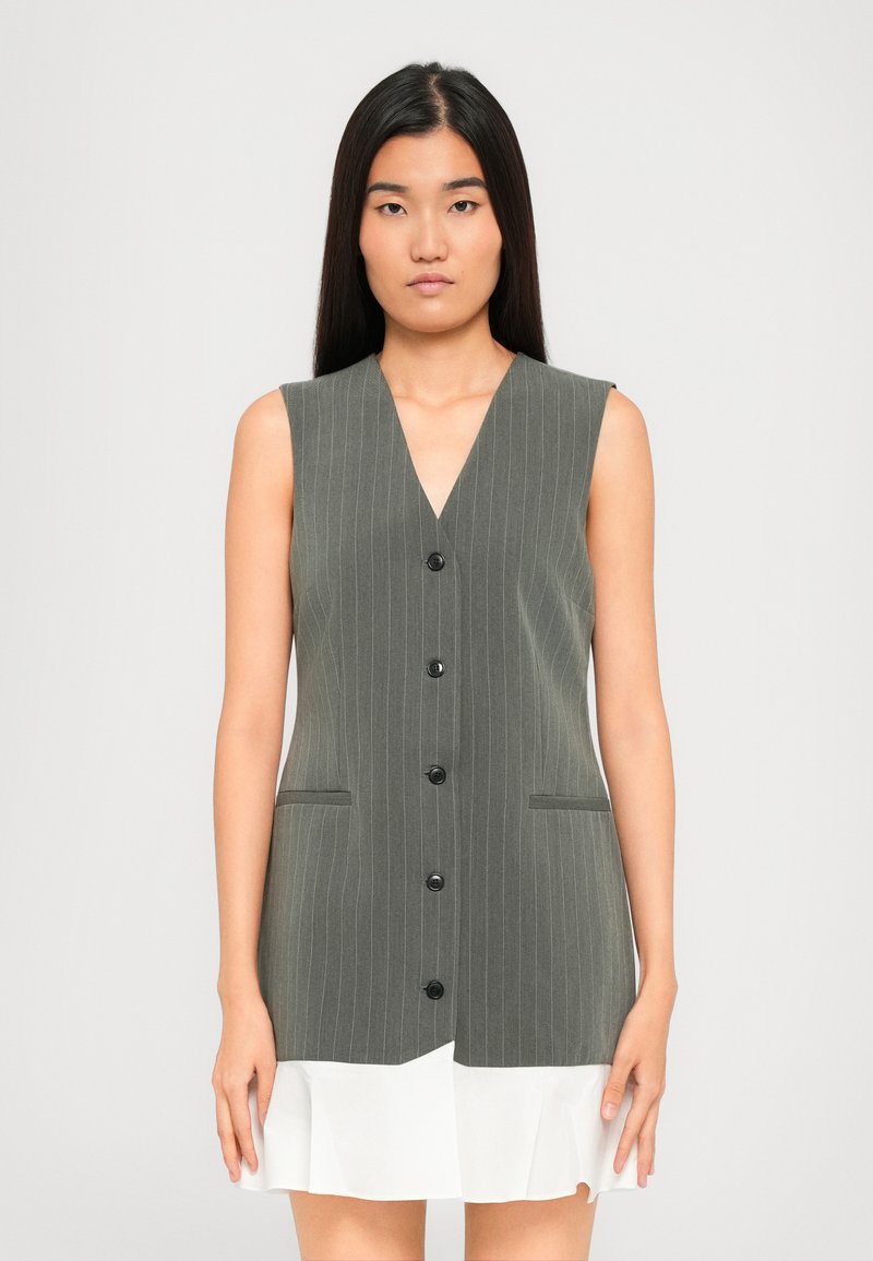 Donna con lunghi capelli neri lisci che indossa un vestito senza maniche a giacca grigia a righe con orlo a volant bianco.