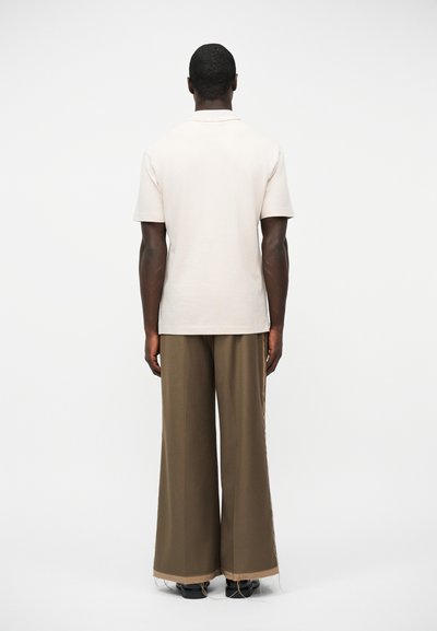 Άνδρας που στέκεται με την πλάτη γυρισμένη, φορώντας ανοιχτό μπεζ κοντομάνικο πουκάμισο και καφέ παντελόνι φαρδιά με σκισμένα τελειώματα και μαύρα παπούτσια.