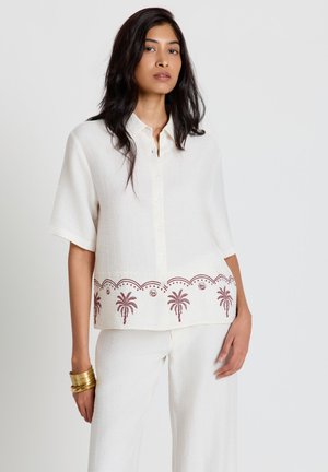 Femme portant une chemise blanche à manches courtes avec une broderie de palmier marron et un pantalon blanc assorti, debout devant un fond uni.