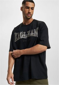 Black cotton t-shirt featuring large white and gold "KARL KANI" graphic print. Short sleeves, relaxed fit, and round neckline.