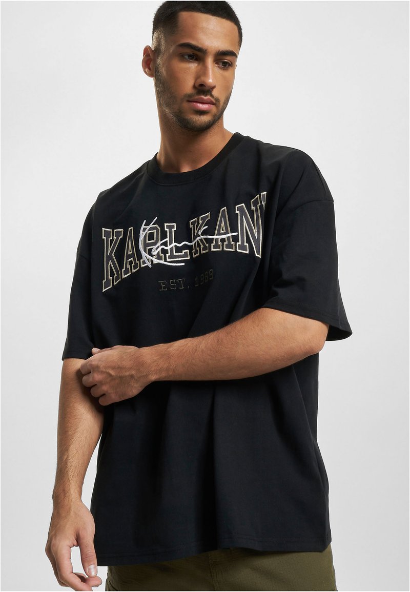 Black cotton t-shirt featuring large white and gold "KARL KANI" graphic print. Short sleeves, relaxed fit, and round neckline.