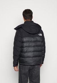 Giacca a vento nera con cappuccio, caratterizzata da un design trapuntato, texture liscia e logo North Face sulla parte superiore della schiena.