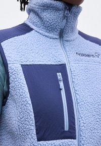 Άτομο που φοράει ανοιχτό μπλε φλις μπουφάν με φερμουάρ στο τσεπάκι του στήθους και το λογότυπο Norrøna στο πάνω μέρος του στήθους.