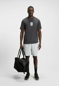 Camiseta gris de manga corta con un logo gráfico, combinada con pantalones cortos blancos. El modelo sostiene un bolso de deporte negro con cremallera. Zapatillas negras con detalles.