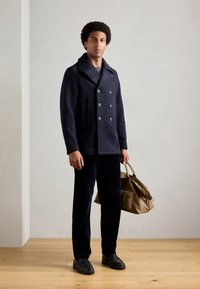 Marineblauer, doppelt geöffneter Peacoat aus Wollmischung mit silbernen Knöpfen; kombiniert mit dunklen Cordhosen und einer braunen Ledertasche.