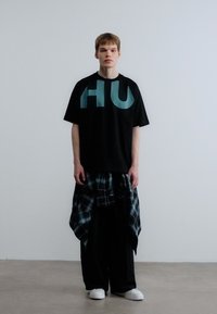 Μαύρο φανελάκι με μεγάλα τιρκουάζ γράμματα "HU", συνδυασμένο με φαρδιά μαύρη παντελόνα και καρό πουκάμισο μαύρο και άσπρο δεμένο στη μέση. Άσπρα παπούτσια.